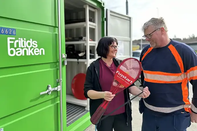 En kvinna håller i ett paket med badmintonracket och tittar leende mot en man. Mannen tittar leende tillbaka. Intill personerna syns en grön yta med texten Fritidsbanken.