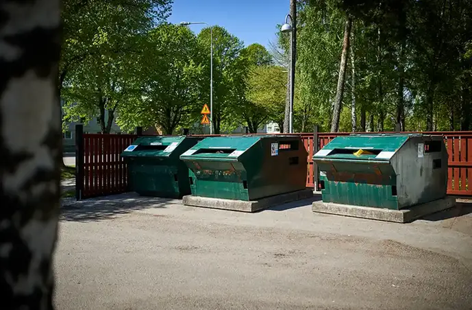 Tre gråna metallcontainrar som omges av ett rött staket. I bakgrunden ses lummiga gröna träd. I förgrunden skymtas en svart och vitt trädstam.